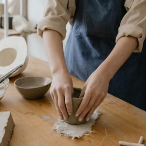 Artisan hands shaping clay on a wooden work table in a pottery studio.