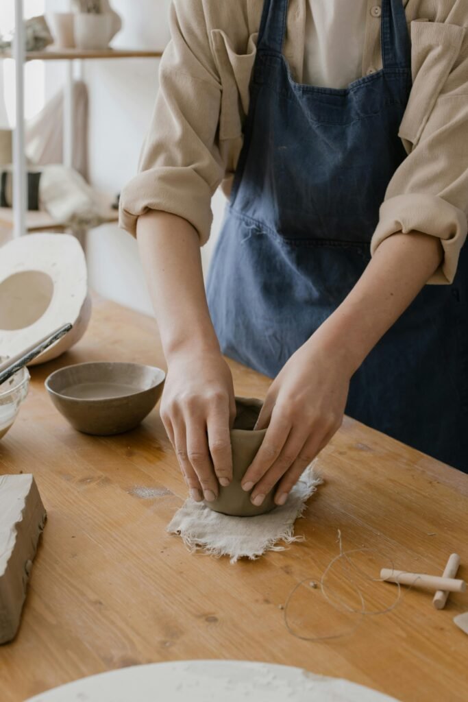 Artisan hands shaping clay on a wooden work table in a pottery studio.