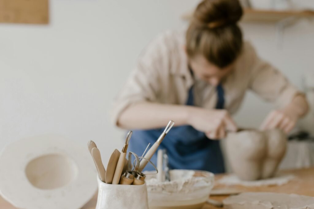 Focused woman sculpting clay in a sunny pottery workshop with tools in the foreground.